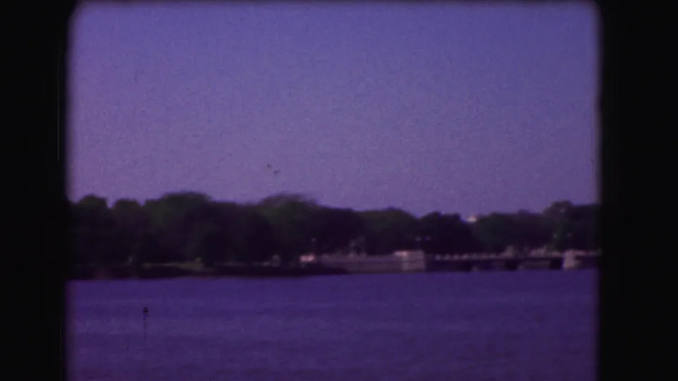 1975 Washington, DC home-movie frame showing open water and a distant wooded shoreline from a boat-like viewpoint.