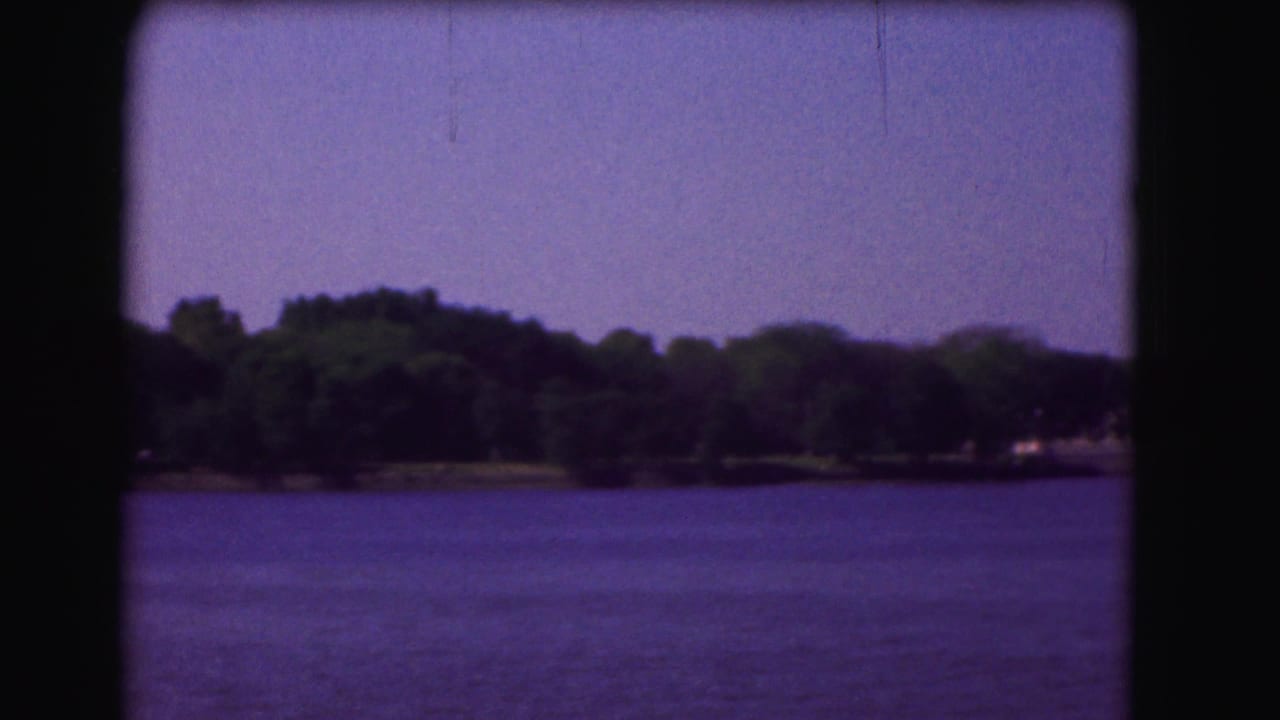 River surface with wooded shoreline and low distant structures beneath a clear sky.
