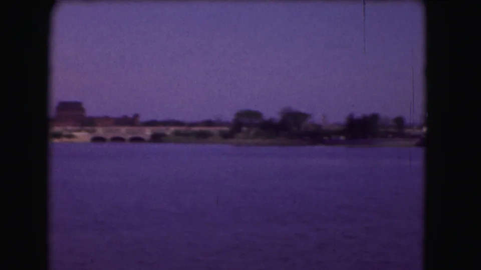 1975 Washington, DC 8mm frame with a low bridge, tree-lined bank, and distant structures across a purple-shifted river view.