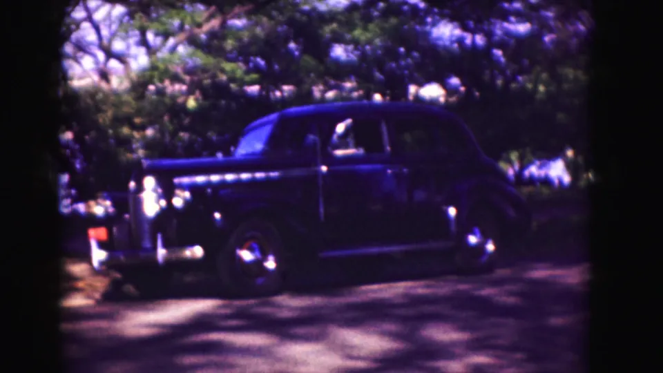 A dark mid-century-style sedan remains parked under trees, with its chrome grille and front fender visible.