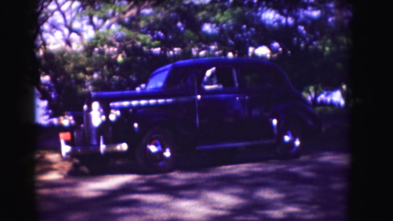 Dark vintage sedan passing trees beside a school-like building.
