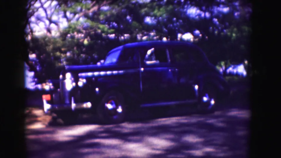 1975 Washington, DC home-movie frame showing a dark sedan near trees and a brick-and-white school-like building.