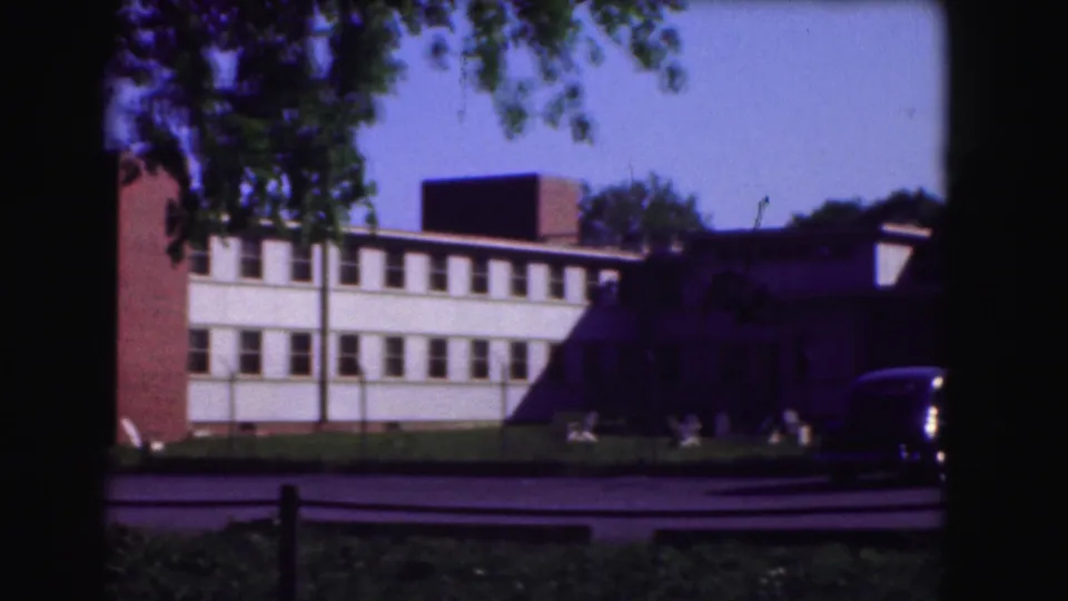 1975 Washington, DC frame of a school-like brick-and-white facade with roadside lawn and a passing sedan partly visible.