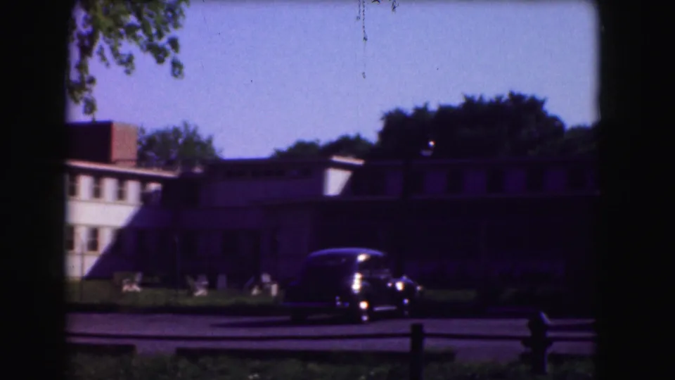 1975 Washington, DC home-movie frame showing a dark sedan on the road in front of a shaded school-like building.