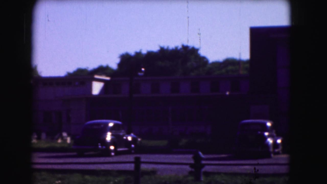 Parked vintage cars outside low buildings, seen through dark window-frame bars.