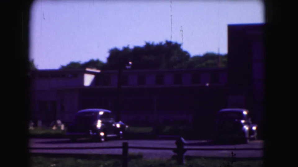 1975 Washington, DC home-movie frame showing parked older cars near low mid-century buildings through a dark foreground frame.