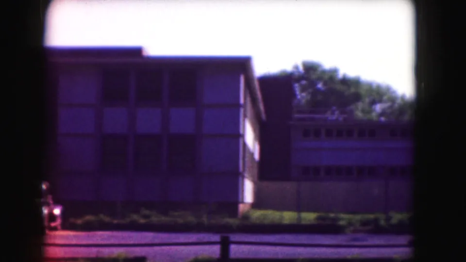 1975 Washington, DC frame showing a sunlit modern building exterior, roadside foreground, shrubs, and a partial parked car.