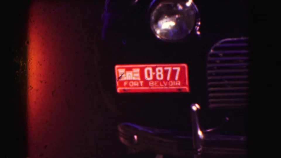 Close 1975 home-movie view of a dark vehicle front with headlight, grille, chrome bumper, and a red Fort Belvoir plate or permit.