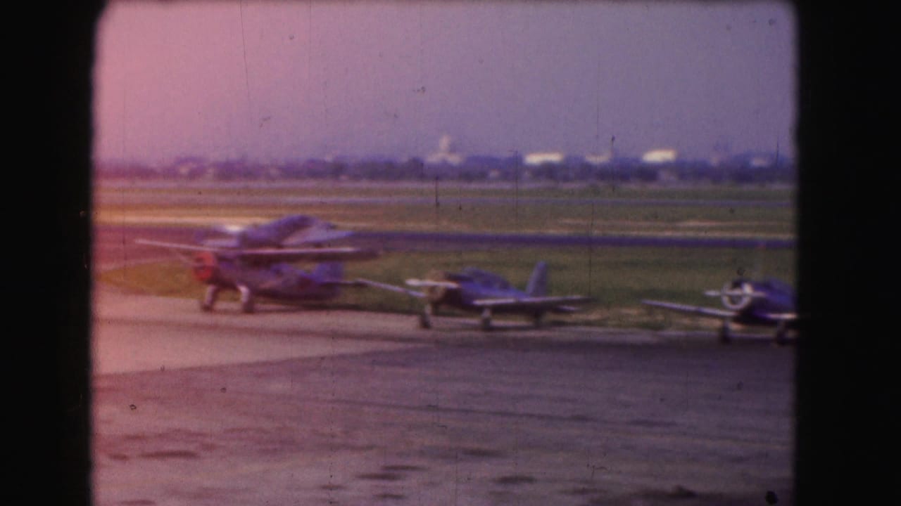 1975 Washington, DC home-movie frame showing several small propeller planes parked on a tarmac beside grassy runway areas.