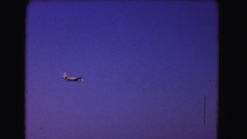 A small airplane passes through the blue-purple sky, captured with soft 8mm grain and visible film wear.