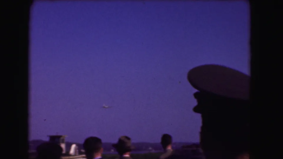 Silhouetted spectators, including a person in a cap, look toward a small plane over the distant airport horizon in Washington DC footage.