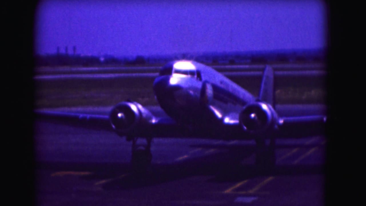 1975 Washington DC home-movie frame showing a shiny twin-engine propeller aircraft taxiing on airport pavement under a blue-purple sky.