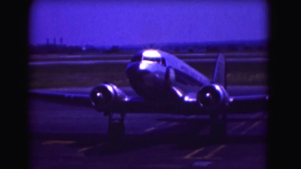1975 Washington DC home-movie frame showing a shiny twin-engine propeller aircraft taxiing on airport pavement under a blue-purple sky.