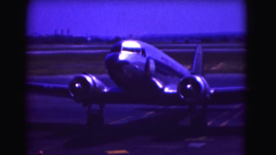1975 Washington DC home-movie still of a twin-engine propeller plane taxiing, with runway markings and distant airfield structures behind it.