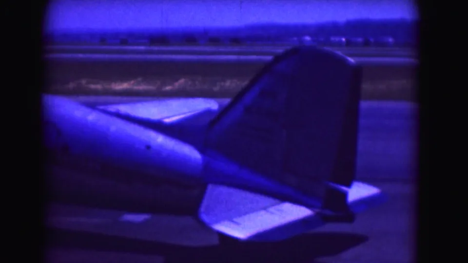 1975 airport home-movie frame showing the rear fuselage and tail of a propeller aircraft on the tarmac in Washington DC.
