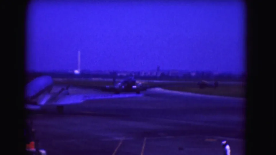 Washington DC airport frame from 1975 showing a distant tarmac view, aircraft tail edge, small figures or vehicles, and runway markings.