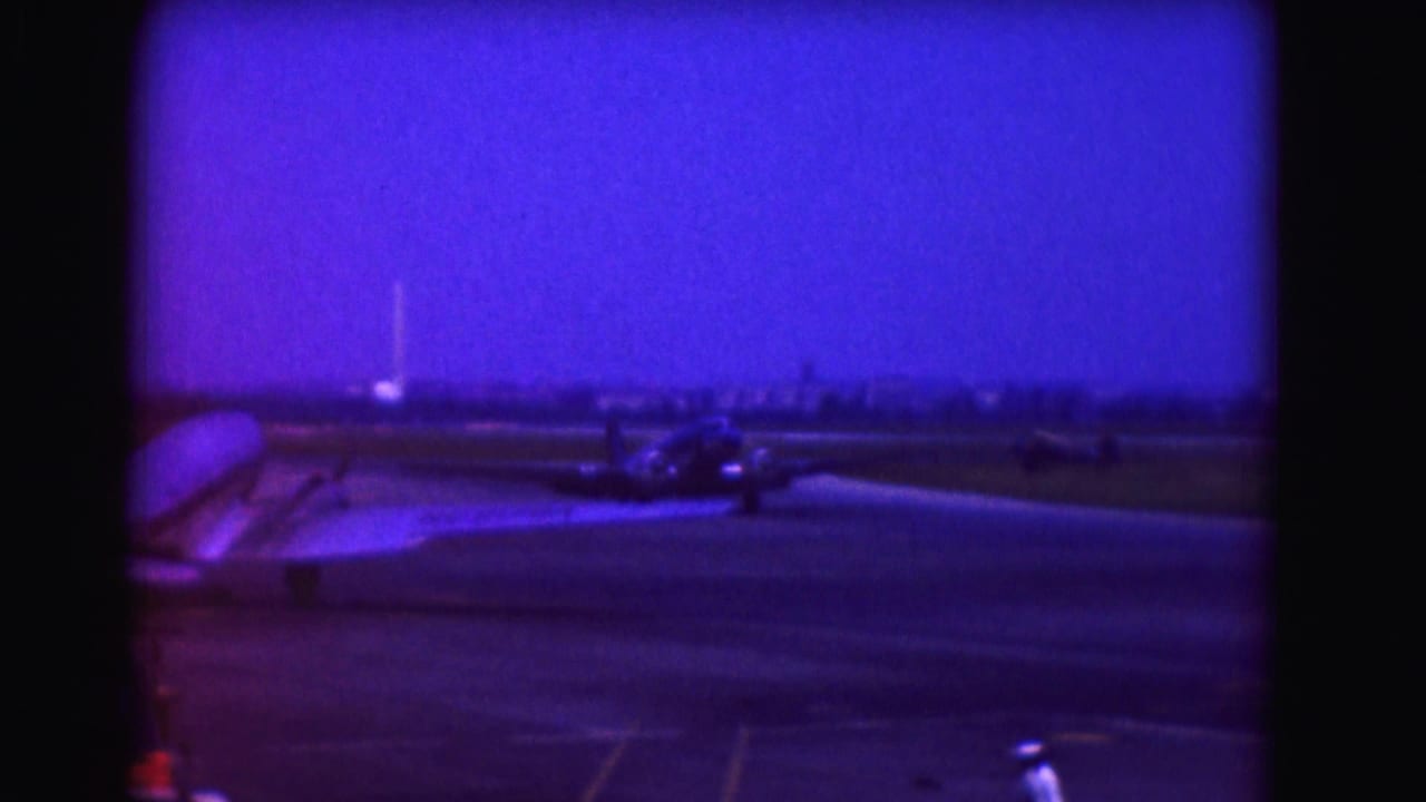 1975 8mm frame of a propeller aircraft on an airport runway, with a distant skyline and a person in white near the lower edge.