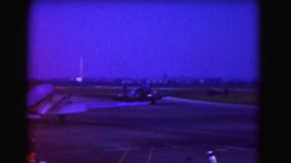 1975 8mm frame of a propeller aircraft on an airport runway, with a distant skyline and a person in white near the lower edge.