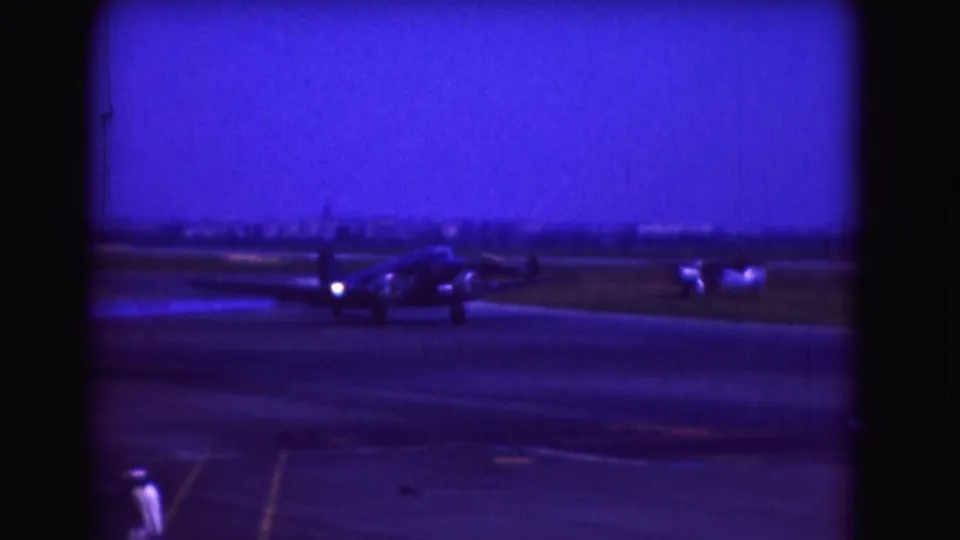Soft 8mm frame of a propeller aircraft taxiing on runway pavement, with a person in white visible at the bottom of the frame.