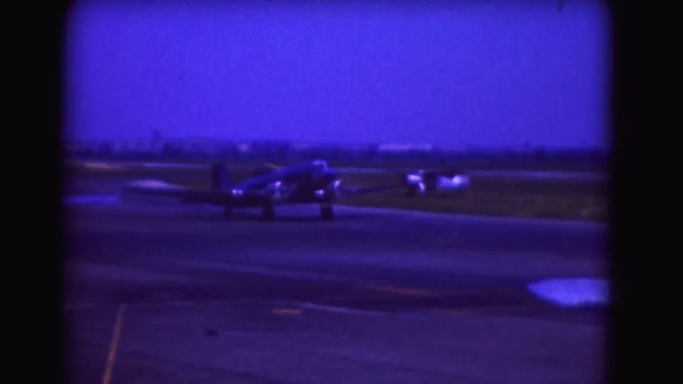 1975 8mm airport frame showing a propeller aircraft taxiing beside runway pavement and grass, with distant structures on the horizon.