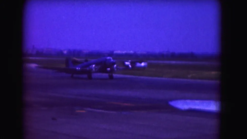 1975 Washington, DC home-movie frame showing a propeller airplane moving along a runway with a distant horizon.