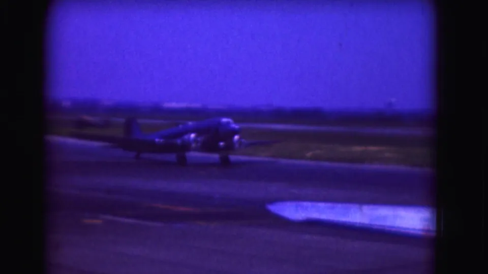 Blue-shifted 8mm frame of the propeller airplane farther along the runway, with open airport ground behind it.