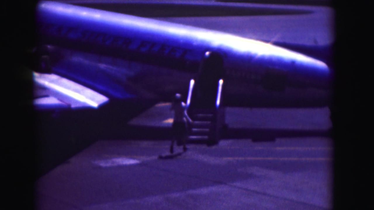 1975 Washington DC home-movie frame showing a passenger running toward portable stairs beside a Pan Am jet under blue night lighting.