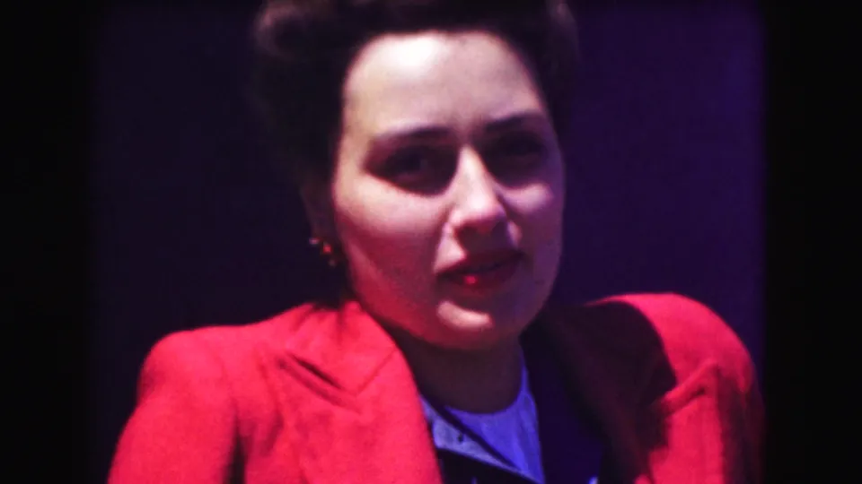 1975 Washington DC home-movie close-up of a woman with short dark hair wearing a red blazer and smiling near a wall or doorway.