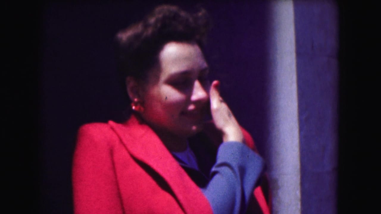 Woman in a red jacket and blue shirt stands beside a column with her hand near her face.
