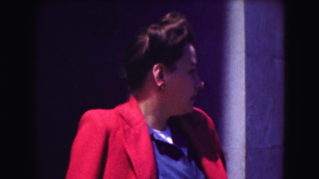 Woman in a red jacket turns in profile beside a column against a dark background.