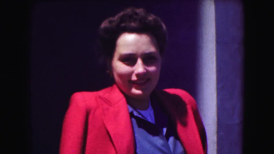 A woman in a red jacket and blue shirt smiles toward the camera near a column in 1975 Washington, DC home-movie footage.