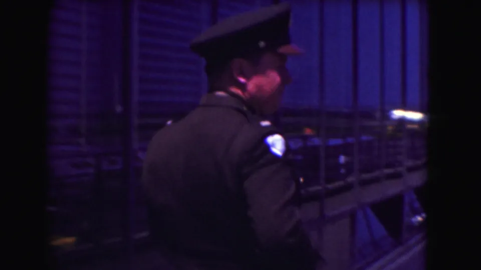 A uniformed police officer appears near a railing and vertical slats in the same 1975 Washington, DC home-movie clip.