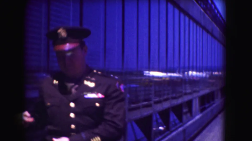 Washington, DC 1975 frame of the officer looking down while standing beside railings and reflective wall panels on a blue-lit platform.