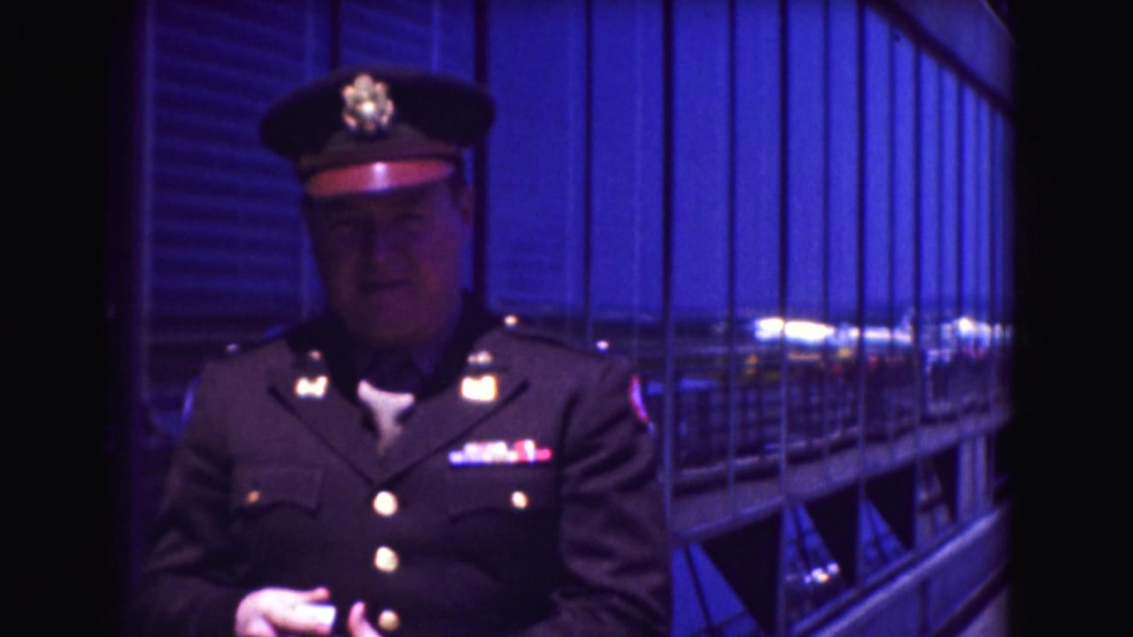 Uniformed man in a peaked cap stands near blue-tinted windows.