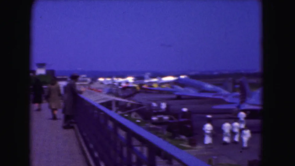 1975 Washington DC home-movie frame showing civilians on an elevated deck overlooking parked aircraft and white-uniformed ground crew.
