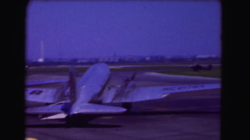 1975 Washington DC airport frame with parked aircraft tails and wings, soft 8mm color, and a distant runway under a blue sky.