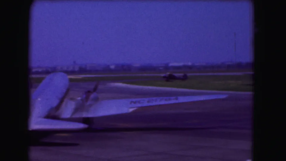 1975 Washington DC home-movie frame showing part of a large propeller airplane on a tarmac, with runway grass and dark vertical framing.
