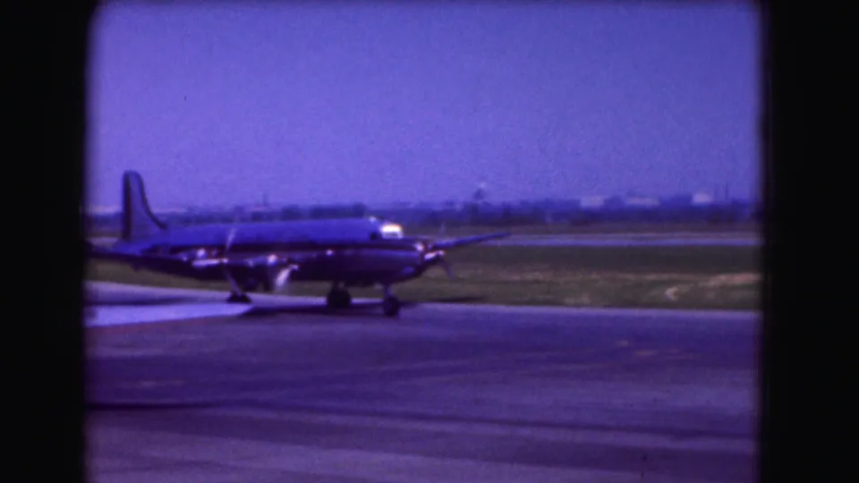 1975 Washington DC frame of a large propeller-driven airplane on airport pavement, with distant structures and a blue-purple 8mm color cast.