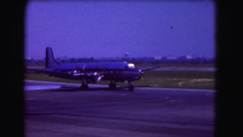 Large propeller aircraft on a Washington DC airport tarmac in 1975 home-movie footage, filmed with soft grain and blue-magenta fading.