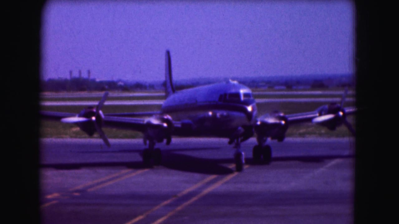 1975 Washington DC home-movie frame showing a silver four-propeller passenger plane moving slowly on airport tarmac.