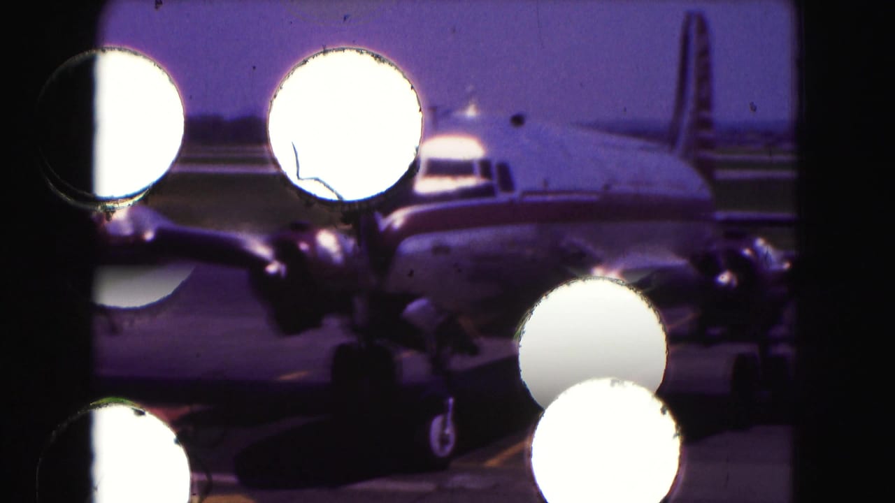 1975 8mm frame of a propeller airplane on a tarmac, partially obscured by bright circular foreground reflections.