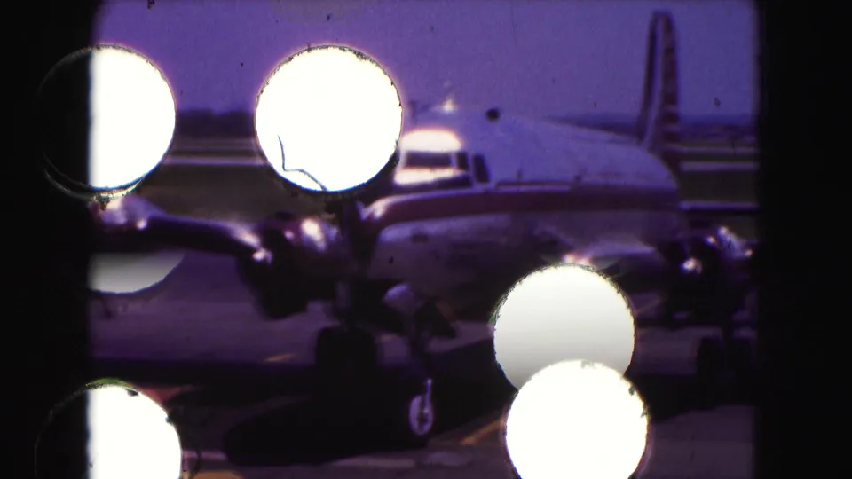 1975 8mm frame of a propeller airplane on a tarmac, partially obscured by bright circular foreground reflections.