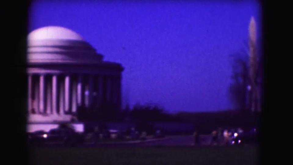 Soft 8mm frame showing part of the Jefferson Memorial, the distant Washington Monument, roadside cars, and open lawn at dusk.