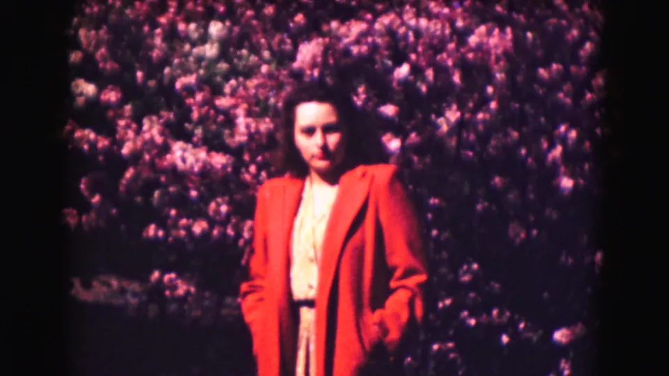 Woman in a red coat poses before pink and purple flowering shrubs in 1975 Washington DC home-movie footage.