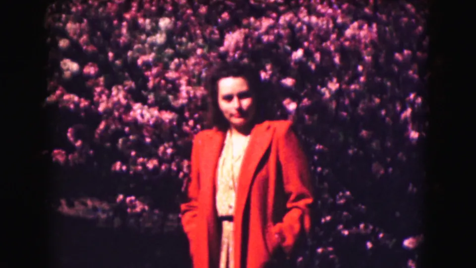 Adult woman in a red coat and light dress stands near flowering shrubs in 1975 Washington DC footage.