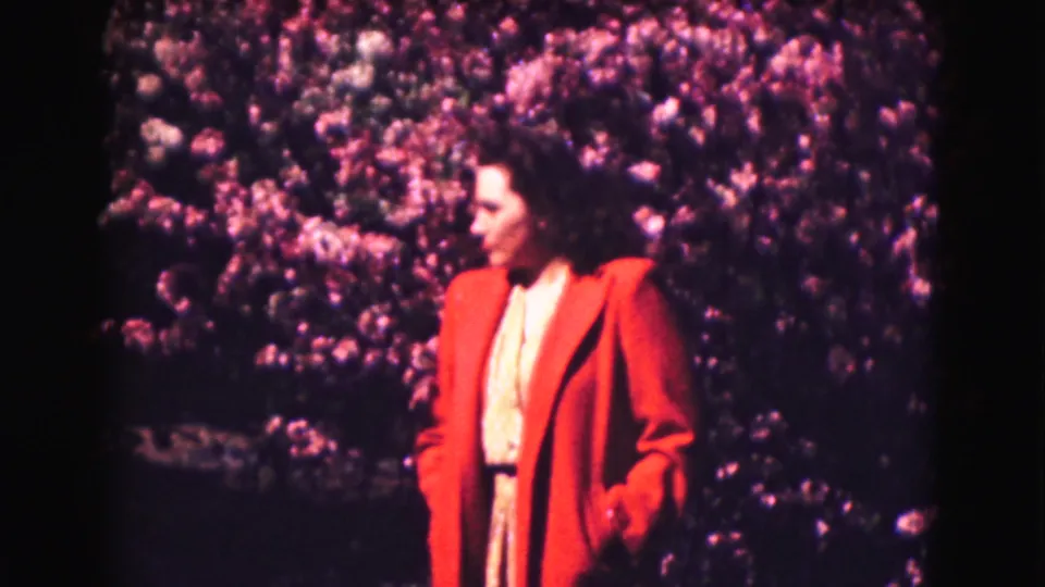 Woman in a red coat turns to the side beside blooming shrubs in 1975 Washington DC home-movie footage.