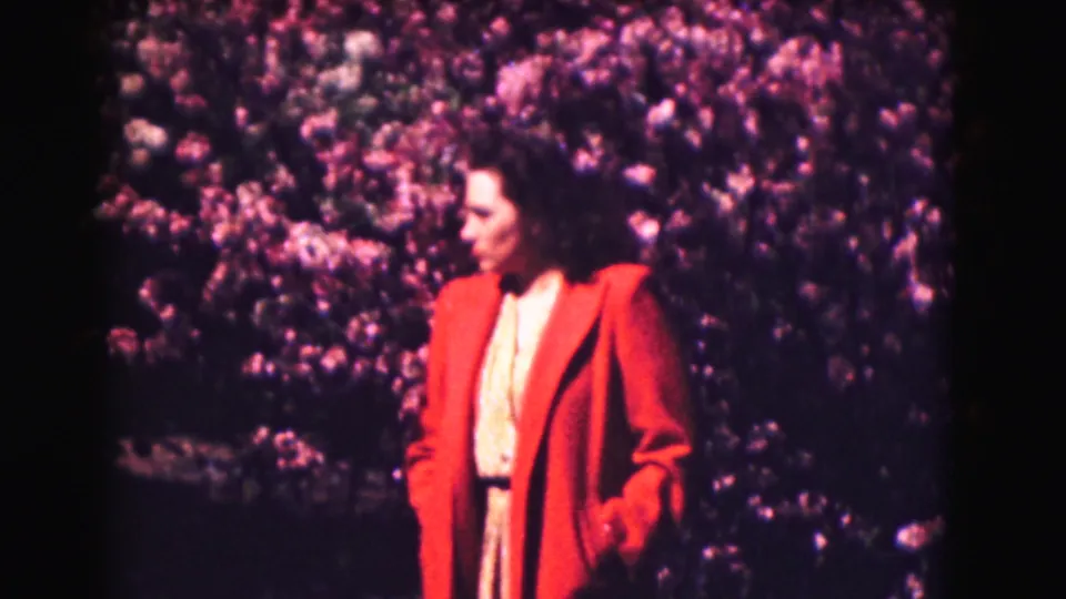 In metadata-supported 1975 Washington DC footage, a woman in a red jacket waits beside a purple-pink flowering hedge.