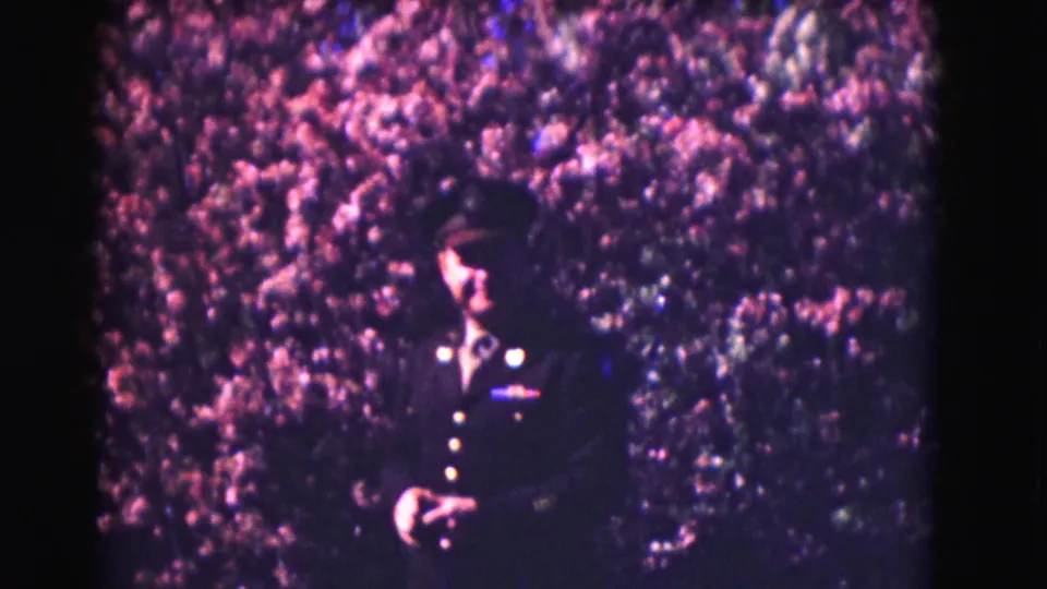 A uniformed officer faces toward the camera near the hedge, with visible 8mm grain and magenta color shift.