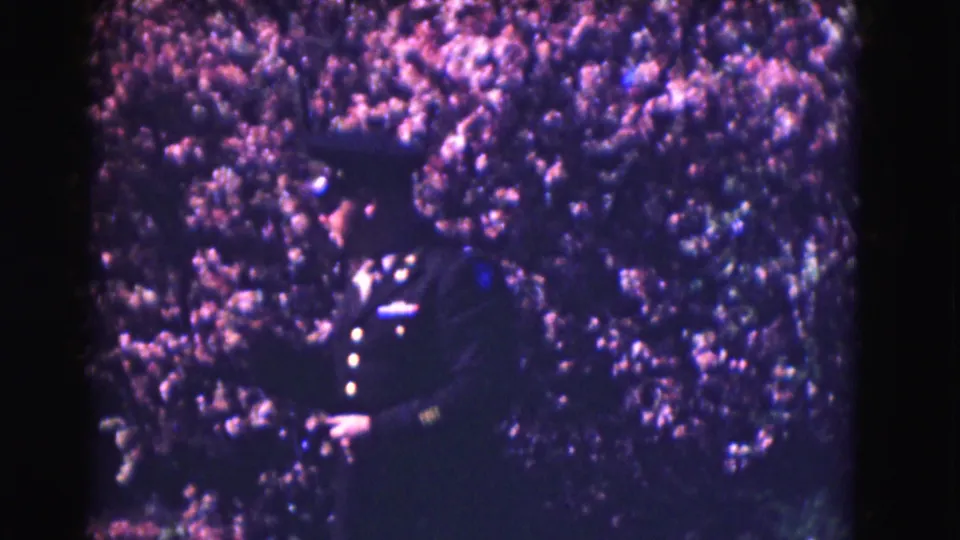 A dark-uniformed officer appears close to the flowering branches, with the home-movie frame soft and grainy.