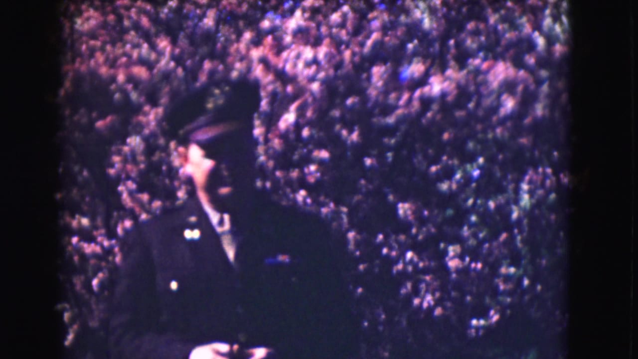 Uniformed man in dark cap standing before dense flowering shrubs.